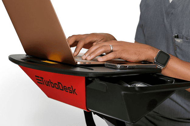 Close-up of a person's hands typing on a laptop that is placed on a mobile desk with a red and black color scheme, branded 'TurboDesk'. The individual is wearing a gray shirt and a smartwatch on their left wrist, which is resting near the laptop's trackpad.