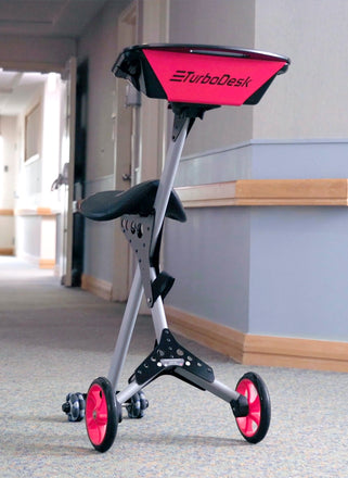 An ergonomic kneeling chair with "TurboDesk" branding on its red storage tray, featuring a silver frame, black pads, and red wheels, stands in an indoor corridor.