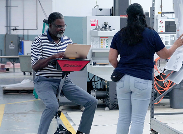 "An indoor industrial setting with a man seated on a Turbo Mobile desk with a red tray, holding a laptop. He is wearing glasses, a striped polo shirt, and gray pants. In front of him stands a woman with her back to the camera, wearing a dark blue shirt and light blue jeans, working on a machine with cables.