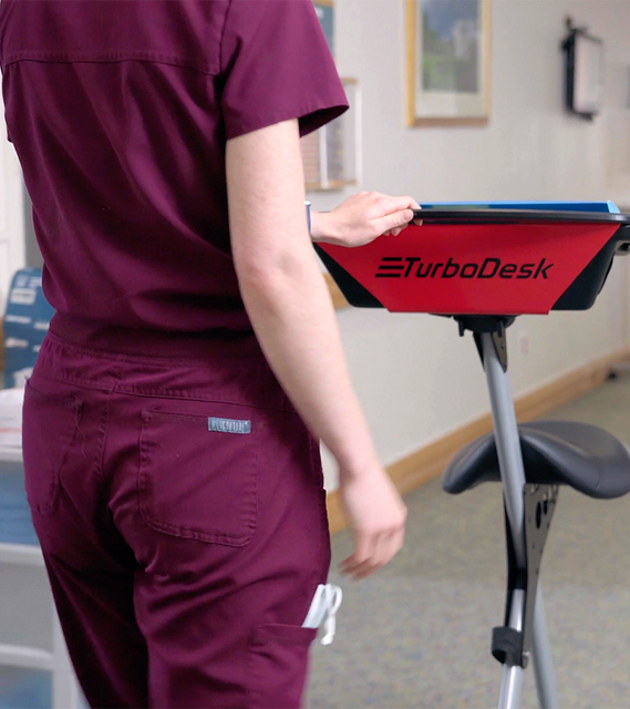 Partial view of a healthcare professional in burgundy scrubs standing beside a mobile workstation with a red tray branded 'TurboDesk'. Only the person's torso and arm are visible as they interact with the workstation. 