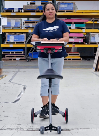 A woman in a blue t-shirt and grey pants stands behind a wheeled ergonomic kneeling chair, holding a clipboard in a warehouse setting.