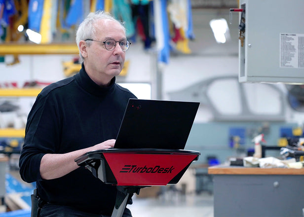 A man in a black turtleneck and glasses stands using a mobile stand-up desk in an industrial workspace. The desk has a red tray with the logo 'TurboDesk' and supports an open laptop. He appears focused on something in the distance, possibly a monitor or another person. 